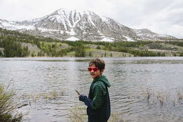 Porträt eines Jungen mit Sonnenbrille, der am See vor den Bergen des Inyo National Forest steht - CAVF41885