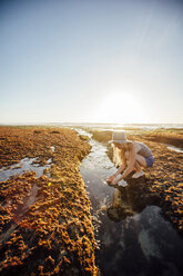 Frau spielt mit Wasser, während sie am Strand gegen den klaren Himmel hockt - CAVF41889