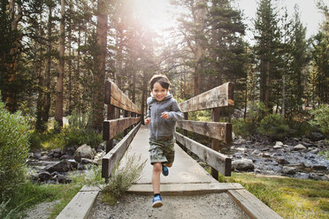 Portrait of boy running over wooden bridge against trees in forest - CAVF42051