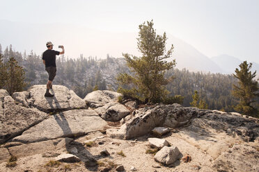 Rückansicht eines Mannes, der auf einem Felsen steht und an einem sonnigen Tag die Berge fotografiert - CAVF42054