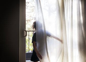Rückansicht eines Mädchens, das vor einem Vorhang am Fenster steht - CAVF42067