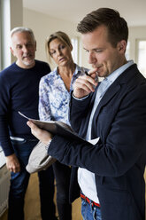 Male real estate agent reading agreement with couple standing in background at home - MASF04892