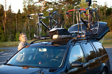 Lächelnde Frau, die neben einem geparkten Auto mit Mountainbikes auf dem Dach an der Straße steht - CAVF42193