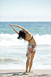 Rear view of woman stretching while standing at sea shore against sky - CAVF42229