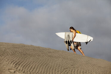 Frau in voller Länge, die ein Surfbrett trägt, während sie auf Sand gegen den Himmel läuft - CAVF42248