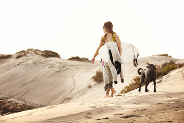Woman carrying surfboard while walking with dog on sand against clear sky - CAVF42268
