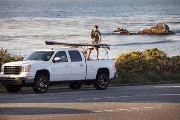 Man tying surfboard on pick-up truck parked at roadside against sea - CAVF42286