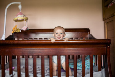 Portrait of happy baby girl standing in crib against wall - CAVF42383