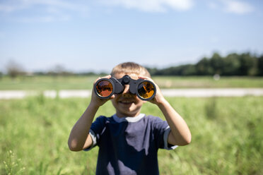 Playful boy looking through binoculars while standing on field during sunny day - CAVF42411