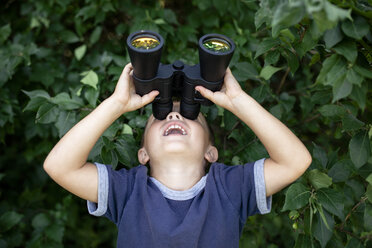 Playful boy looking through binoculars while standing against plants - CAVF42413