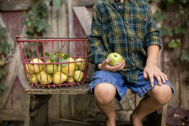 Midsection of boy holding pear while sitting by basket against cottage - CAVF42436