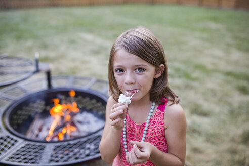 Hochwinkliges Porträt eines Mädchens, das geröstete Marshmallows isst, während es im Hof mit einer Feuerstelle im Hintergrund steht - CAVF42456