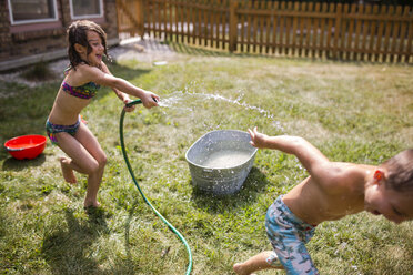 Playful sister spraying water on shirtless brother through hose at yard - CAVF42461