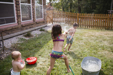 Baby girl looking at sister spraying water on shirtless brother through hose - CAVF42462