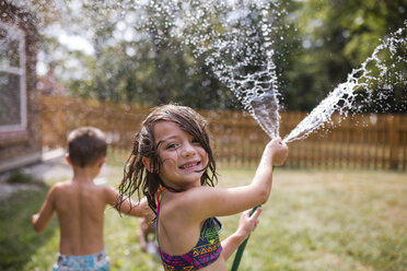 Portrait of sister spraying water through garden hose with shirtless brother standing in background - CAVF42463