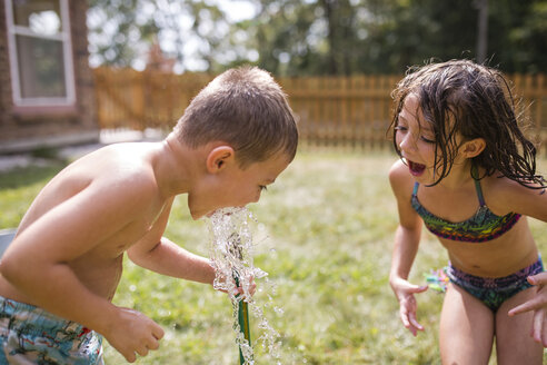 Schwester mit offenem Mund, die ihren Bruder beim Trinken von Wasser durch einen Gartenschlauch im Garten beobachtet - CAVF42464