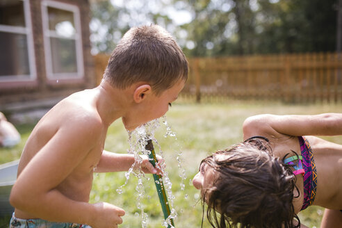 Verspielte Schwester, die ihren Bruder beim Trinken von Wasser aus dem Gartenschlauch beobachtet, während sie sich im Garten bückt - CAVF42465