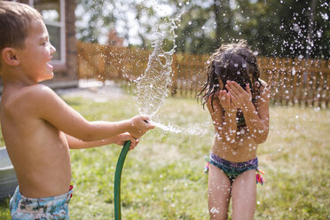 Shirtless brother spraying water on sister through garden hose at yard - CAVF42466