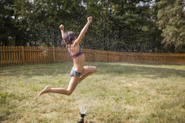 Cheerful girl jumping over sprinkler at yard - CAVF42467