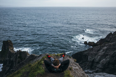 High angle view of hiker sitting with hands behind head on cliff against seascape - CAVF42482