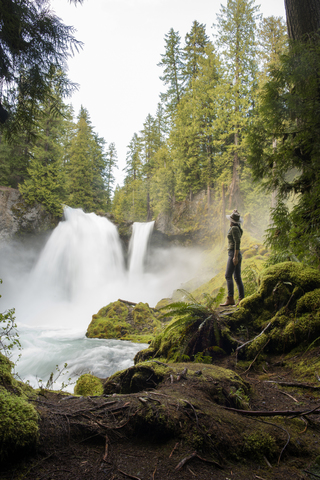 Seitenansicht einer Wanderin, die auf einem moosbewachsenen Felsen stehend einen Wasserfall betrachtet, lizenzfreies Stockfoto