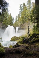 Seitenansicht einer Wanderin, die auf einem moosbewachsenen Felsen stehend einen Wasserfall betrachtet - CAVF42502