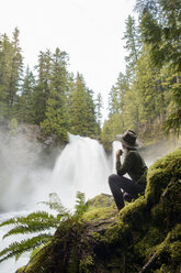 Seitenansicht einer Wanderin, die auf einem moosbewachsenen Felsen sitzend einen Wasserfall betrachtet - CAVF42503