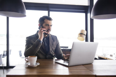 Businessman using laptop and talking on smart phone while sitting table in cafe - CAVF42604