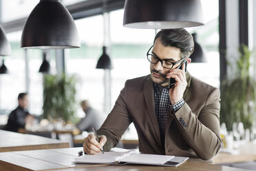 Businessman writing in diary while talking on smart phone at table in restaurant - CAVF42653