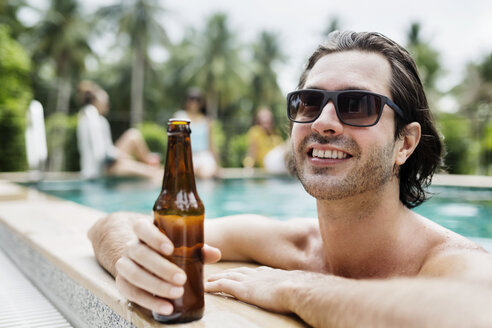 Thoughtful man holding beer bottle in swimming pool - CAVF42717