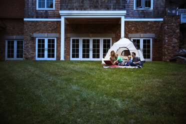 Familie isst Pizza und sitzt beim Zelt auf der Wiese vor dem Haus - CAVF42824