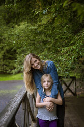 Portrait of smiling mother and daughter standing by railing against plants in yard - CAVF42829