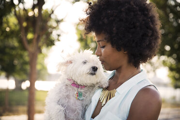 Woman with curly hair kissing dog while standing at park - CAVF42860