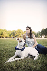 Nachdenkliche Frau sitzt mit Hund auf einer Wiese im Park gegen den klaren Himmel - CAVF42866