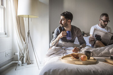Schwuler Mann frühstückt, während sein Partner zu Hause auf dem Bett Zeitung liest - CAVF43005
