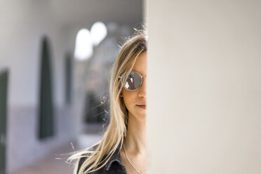 Portrait of young woman wearing mirrored sunglasses hiding behind column - AFVF00440