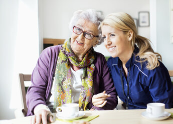 Happy female caretaker and senior woman looking away while sitting at table in nursing home - MASF04945