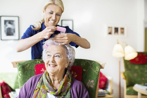 Happy female caretaker putting curlers to senior woman's hair at nursing home stock photo