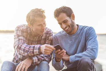 Glückliche männliche Freunde, die eine Textnachricht auf einem Mobiltelefon am Meer lesen - MASF04961