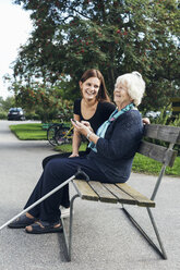 Glückliche Großmutter und Enkelin benutzen ein Mobiltelefon auf einer Parkbank - MASF04988