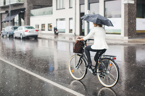 Full length rear view of businesswoman riding bicycle on wet city street during rainy season stock photo