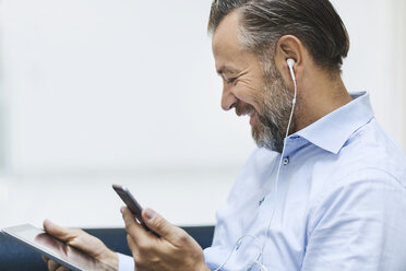 Zufriedener Geschäftsmann, der ein digitales Tablet benutzt, während er über sein Mobiltelefon in der Büro-Lobby Musik hört - MASF05035