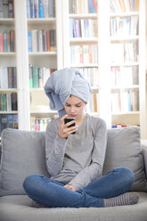 Mädchen mit Handtuch-Turban sitzt zu Hause auf der Couch und schaut auf ihr Handy - LVF06882