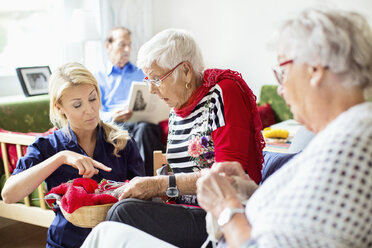 Female caretaker assisting senior women in knitting while man reading book in background at nursing home - MASF05067