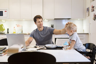 Father stroking baby boy while using laptop at dining table - MASF05078