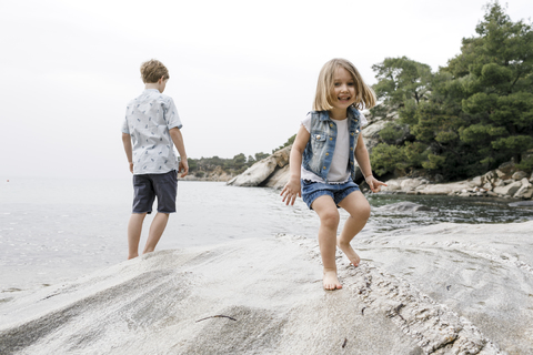 Griechenland, Chalkidiki, Porträt eines glücklichen kleinen Mädchens am Strand mit Bruder im Hintergrund, lizenzfreies Stockfoto
