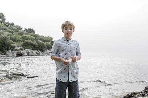 Greece, Chalkidiki, portrait of blond boy standing in front of the sea holding stone stock photo