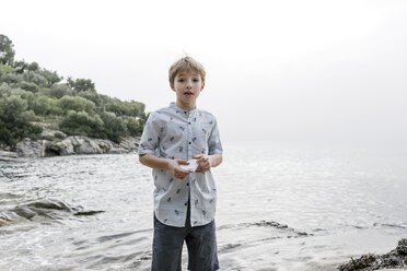 Griechenland, Chalkidiki, Porträt eines blonden Jungen, der einen Stein vor dem Meer hält - KMKF00164