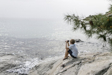 Griechenland, Chalkidiki, Junge mit Fernglas sitzt auf einem Felsen und schaut auf das Meer - KMKF00168