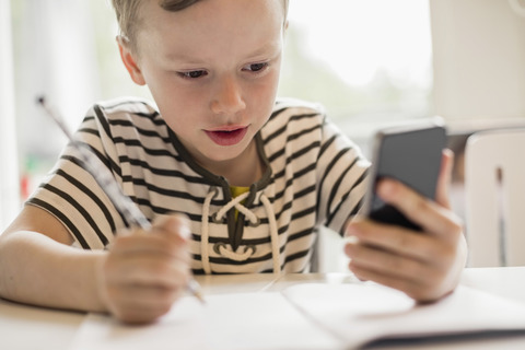 Junge benutzt Smartphone beim Schreiben am Tisch, lizenzfreies Stockfoto